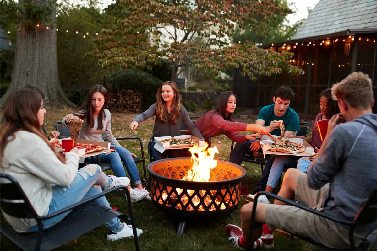 Safety Tips When Using a Fire Pit Image of a group of teenagers eating pizza around a burning fire pit.