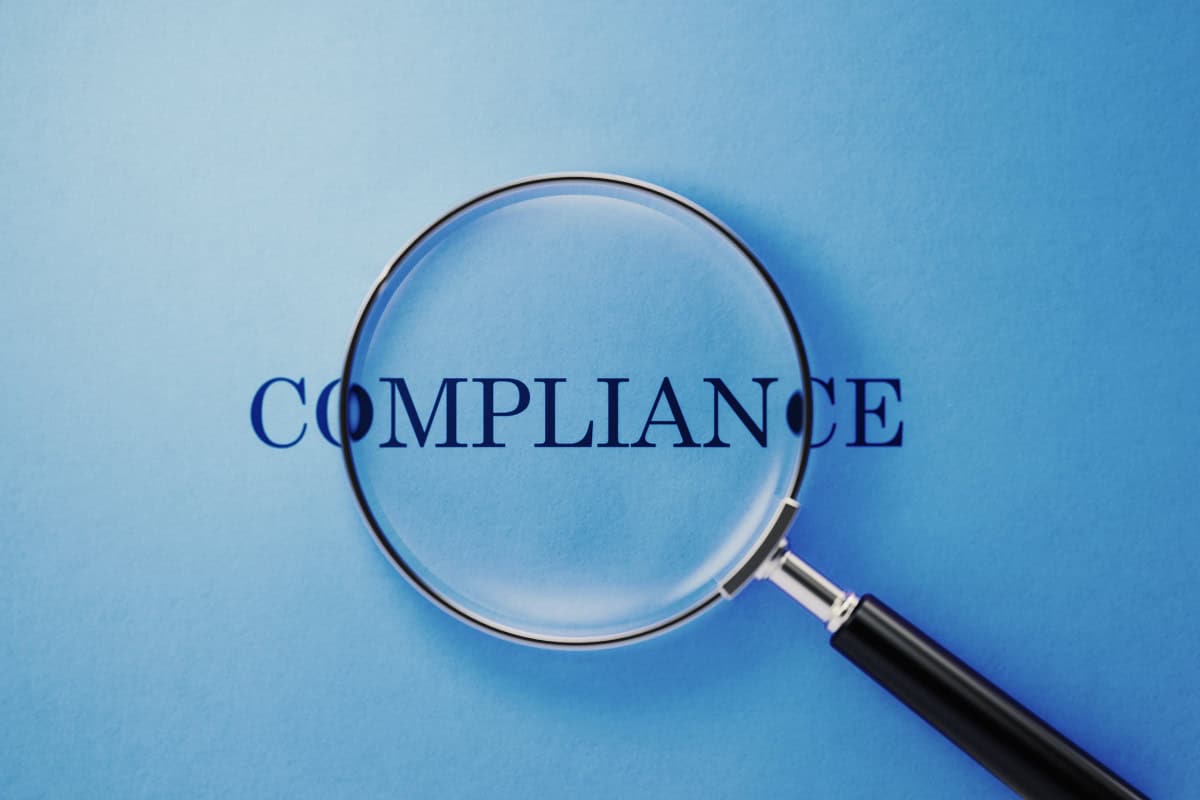 Image of a magnifying glass looking at the word "compliance" in the center of the page.