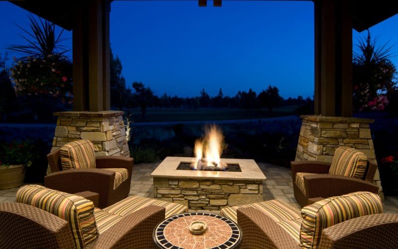Image of an evening scene showing a glowing gas fire pit under covered patio with comfortable outdoor chairs arranged around the flames for cozy nighttime gathering.